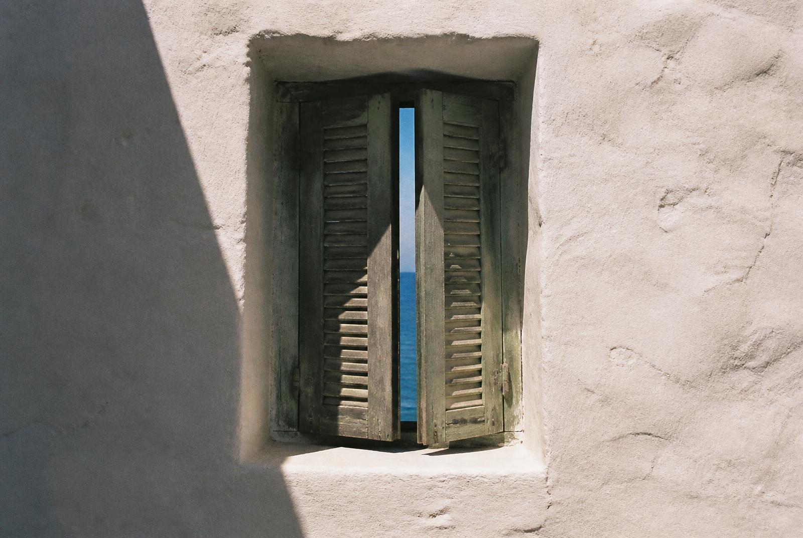 Ventana pequeña de muro grueso encalado con contraventanas de madera y vista al mar